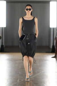 Female model walks a runway in a dark tank top and oversized black bubble skirt, with a belt, sunglasses, and teal flip-flops.