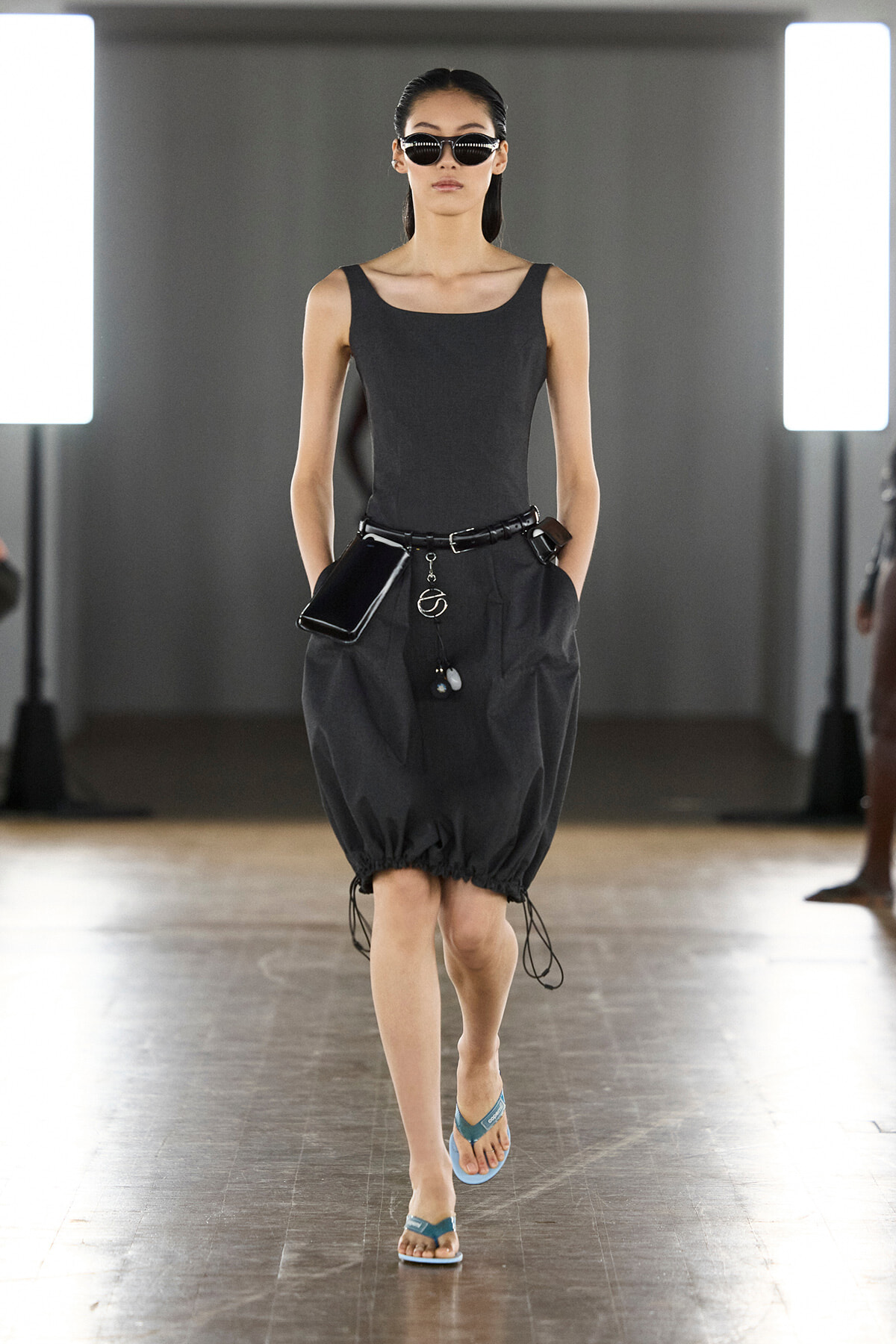 Female model walks a runway in a dark tank top and oversized black bubble skirt, with a belt, sunglasses, and teal flip-flops.
