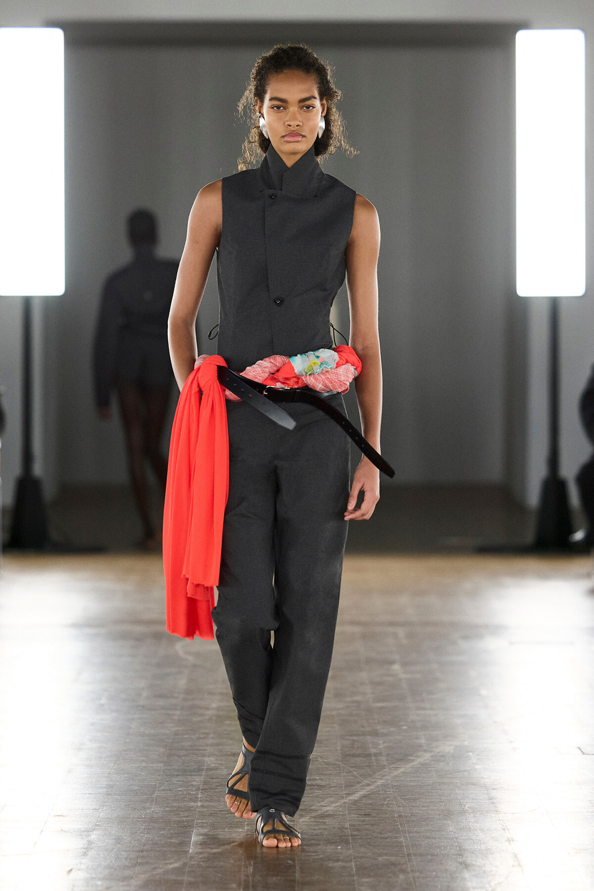 Model walking a fashion runway in a dark sleeveless jumpsuit with a bright red scarf tied around the waist and a black belt, sandals visible.