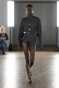 Black model walks a fashion show wearing a charcoal gray romper with oversized cuffed sleeves, a black belt, and a small black pouch at the hip; minimalist jewelry visible.