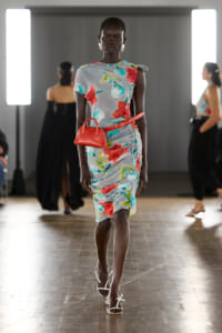 Model walking a runway in a light gray floral dress with red flowers, a red belt bag, and white sandals.