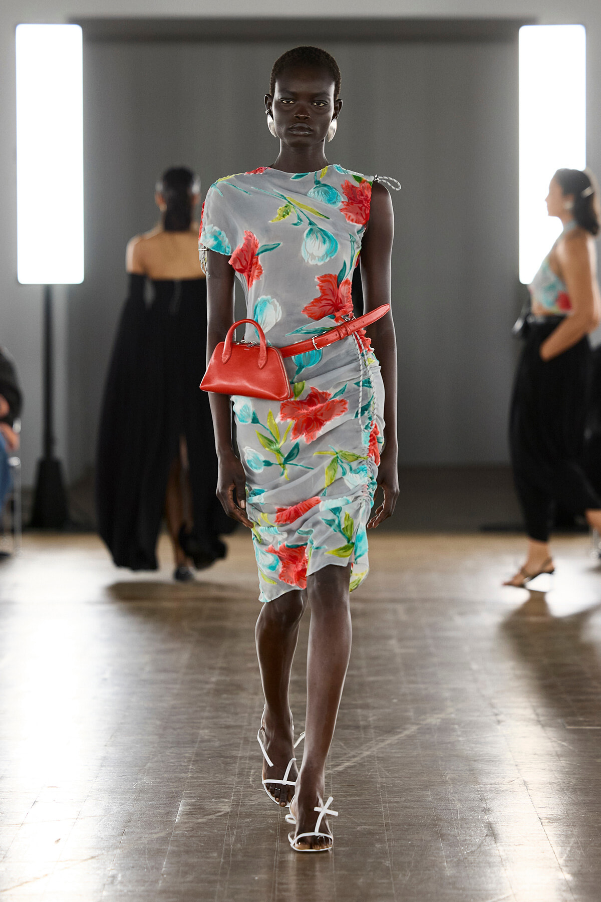Model walking a runway in a light gray floral dress with red flowers, a red belt bag, and white sandals.