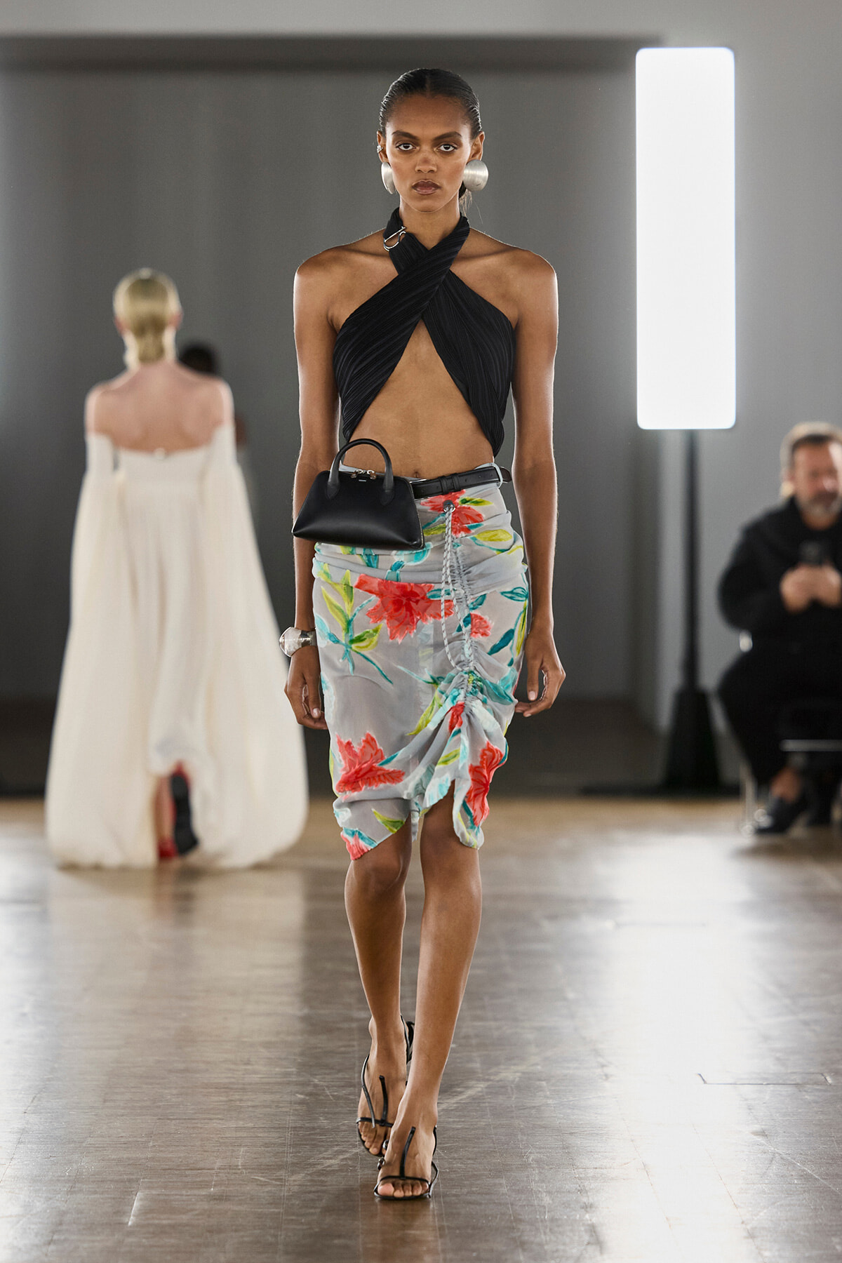 Model walking the runway in a black crisscross halter top, floral knee-length skirt, and strappy heels, carrying a small black handbag.