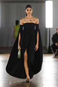 Model walking a runway in a black off-shoulder, floor-length gown with a high-front slit, carrying a green bottle-shaped clutch.