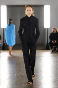 Female model walks a fashion runway in a structured black pinstripe suit with a fitted jacket and wide-leg pants, spotlighted backstage lighting.