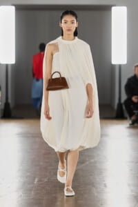 Model walking a runway in a flowing cream halter dress, carrying a small brown handbag and wearing white ballet flats
