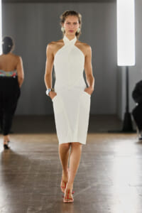Model walks the runway in a white, textured halter-neck midi dress with pockets, pairing with red sandals.