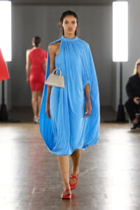 Model walking a fashion show in a bright blue draped cape dress, carrying a small beige handbag.