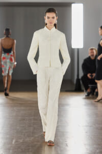 Model walking a runway in a white textured pantsuit with a high collar and vertical seam details.