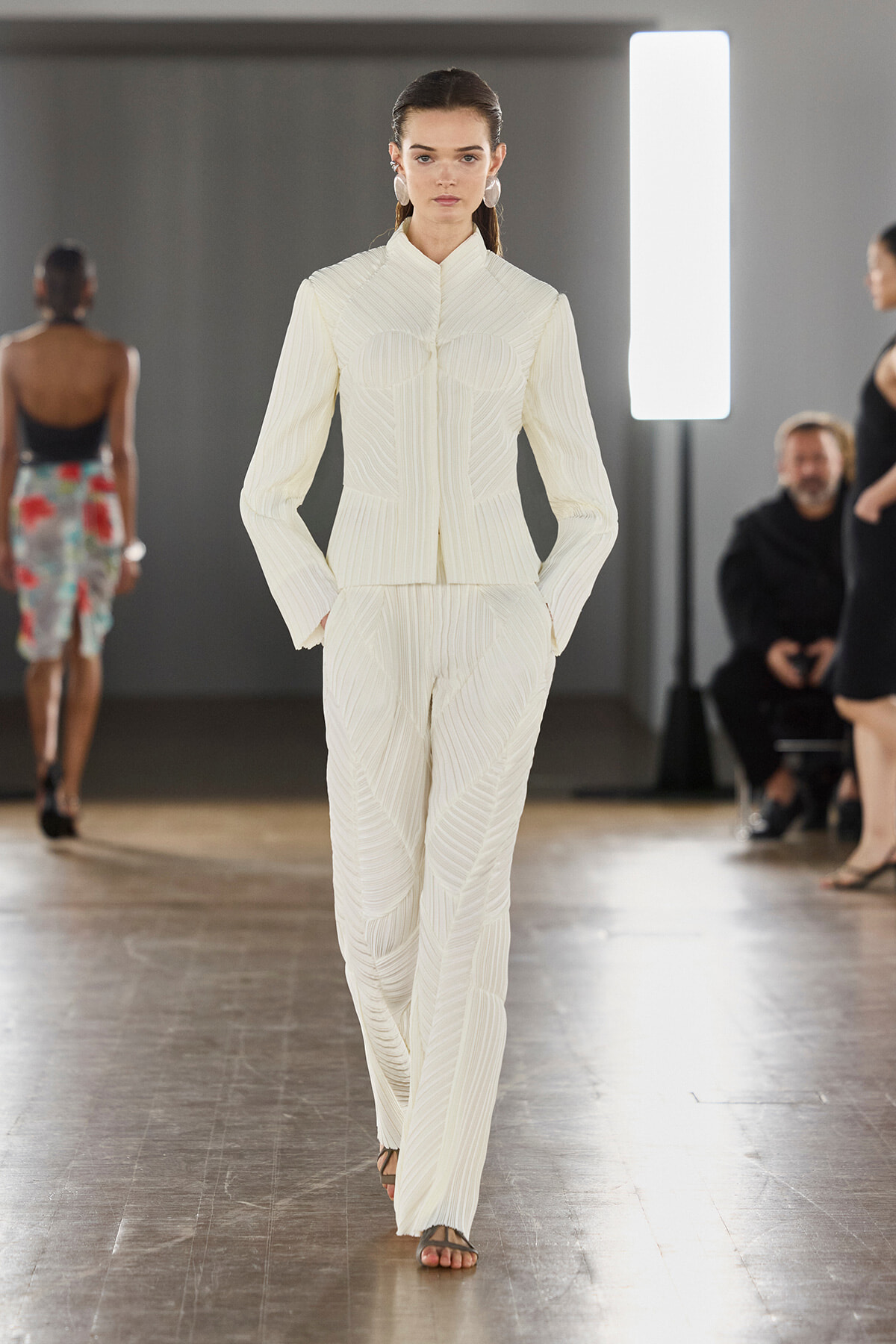 Model walking a runway in a white textured pantsuit with a high collar and vertical seam details.