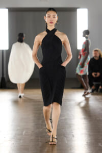 Model walks the runway in a black pleated halter dress with a cross-front design, hands in pockets, wearing strappy sandals.
