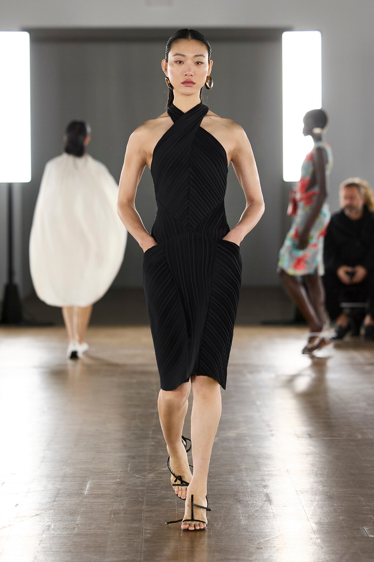 Model walks the runway in a black pleated halter dress with a cross-front design, hands in pockets, wearing strappy sandals.