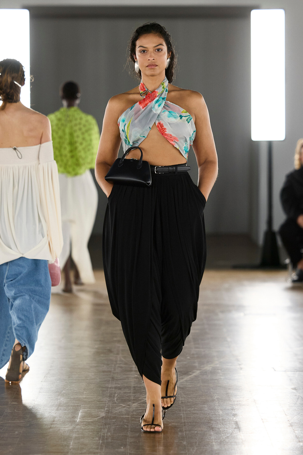 Model walking a runway in a colorful tropical halter top and black draped pants, carrying a small black handbag.