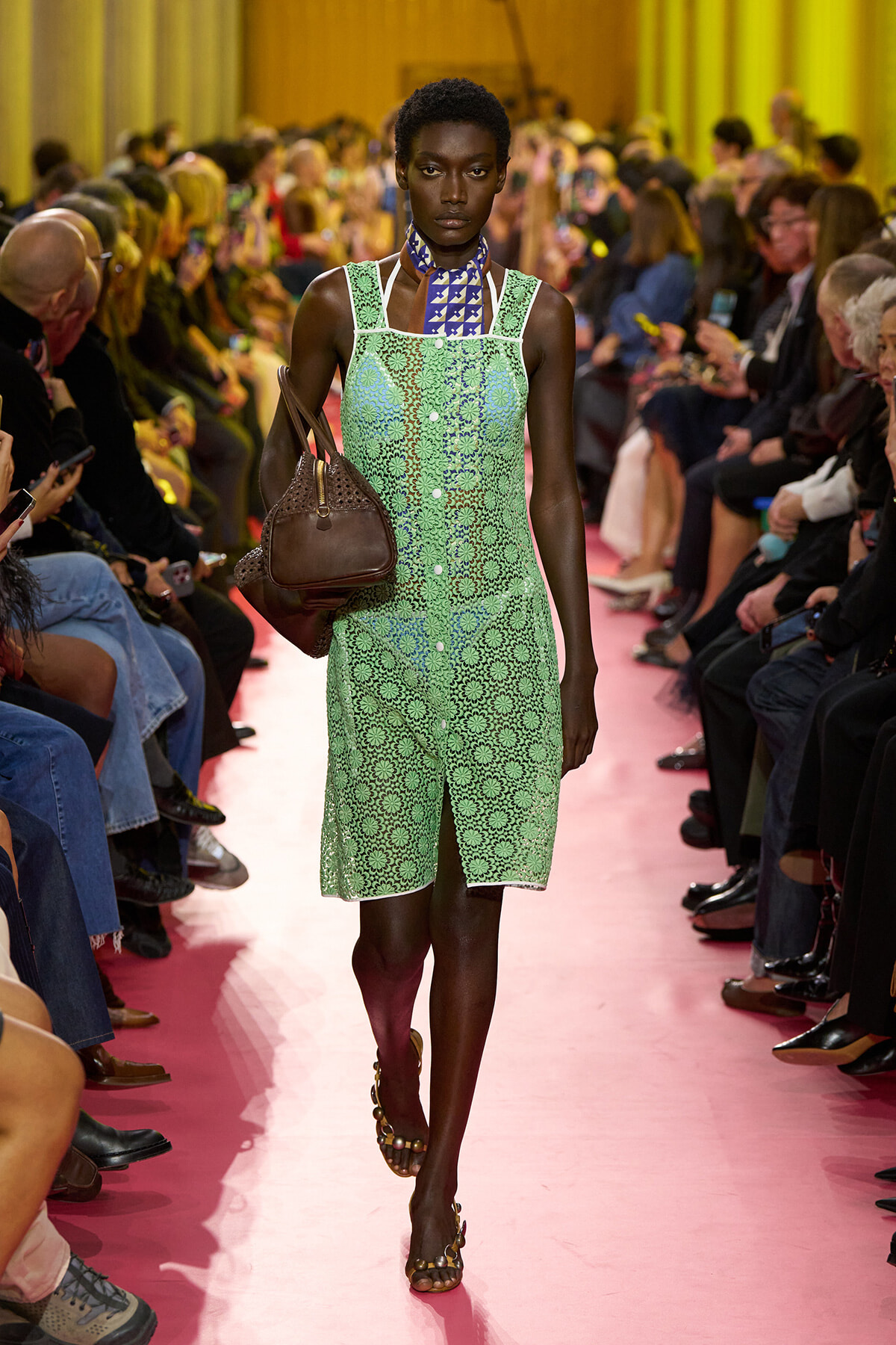 Dark-skinned female model walks a pink fashion-show runway in a mint-green lace dress with a blue patterned neck scarf, carrying a brown handbag as spectators watch.