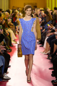 Model walks a pink runway in a blue floral dress with cutouts and ruffled sleeves, carrying a mustard handbag.