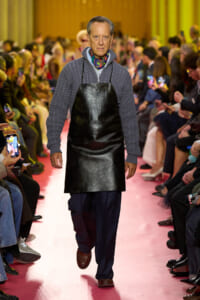 Male model walks a pink runway in a black leather apron over a gray knit sweater, with a colorful scarf nearby audience.