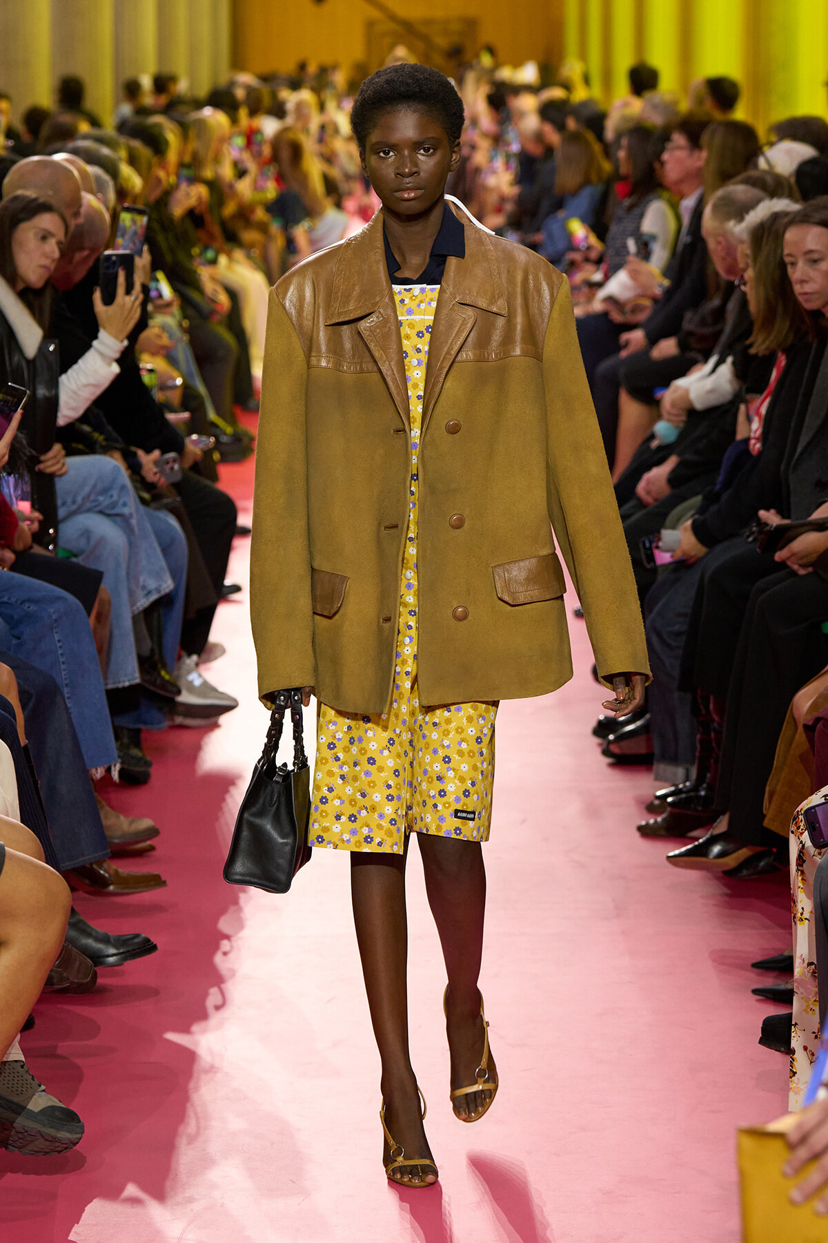 Model walking a pink runway in a mustard-brown coat over a yellow floral dress, audience watching on both sides.