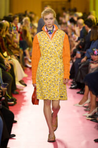 Model walks the runway in a yellow floral wrap dress with orange knit sleeves, pink floor, and a red purse on her left.