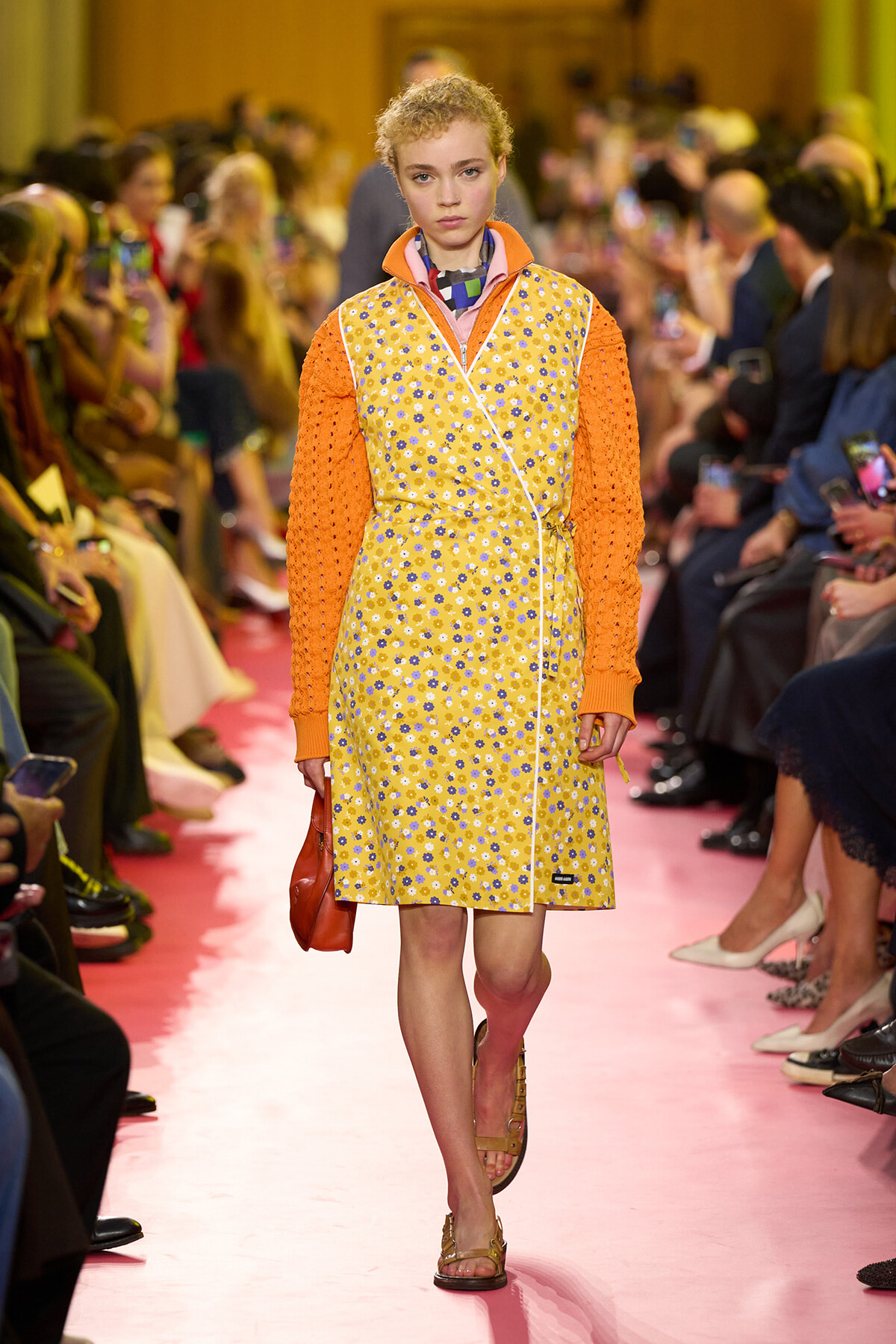 Model walks the runway in a yellow floral wrap dress with orange knit sleeves, pink floor, and a red purse on her left.