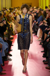 Model walks down a pink runway in a dark blue lace dress, carrying a tan woven bag, surrounded by watching spectators.