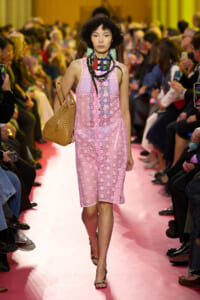 Model walking down a pink runway in a pink lace dress, carrying a tan handbag with a crowd watching the show in the background