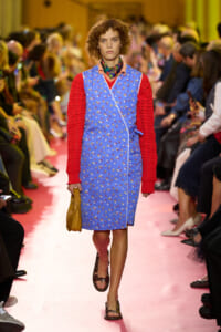 Model walking a pink runway in a blue floral wrap dress with red knit sleeves, tan handbag, and brown sandals at a fashion show.