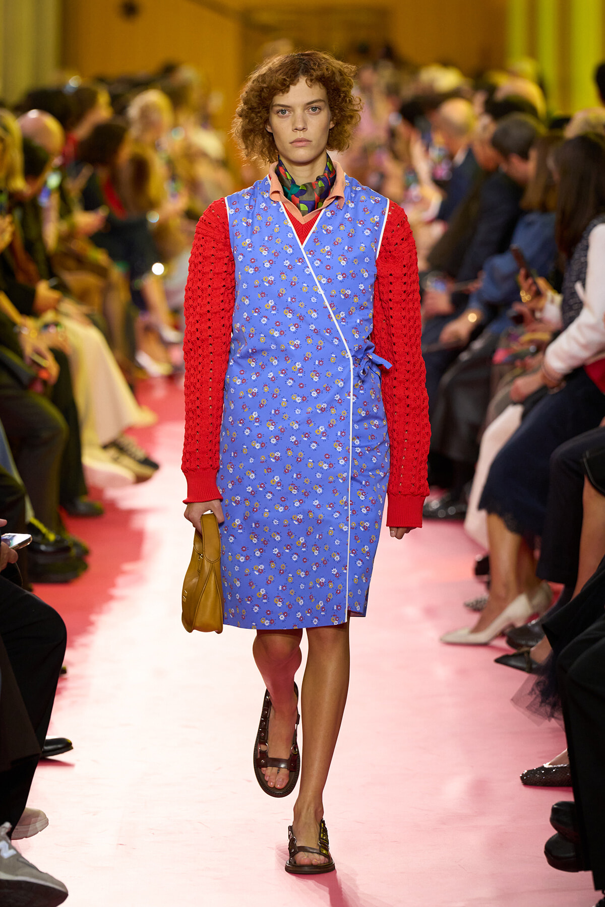 Model walking a pink runway in a blue floral wrap dress with red knit sleeves, tan handbag, and brown sandals at a fashion show.