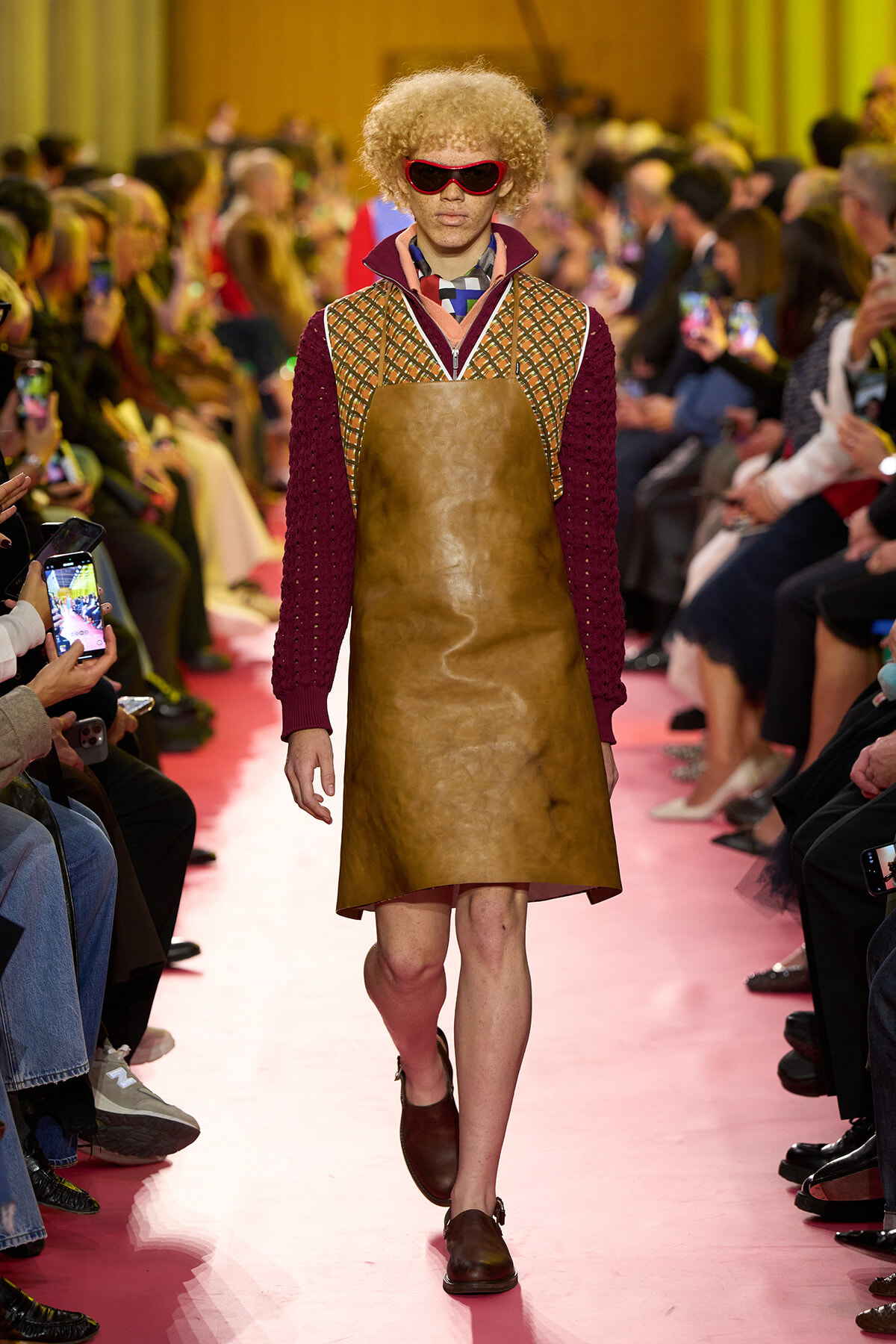 Model with curly blond hair wearing red sunglasses walks the pink runway in a brown leather apron over a burgundy knit sweater and patterned vest.