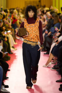 Female model walks a pink runway in an orange floral ruffled top, maroon turtleneck, dark wide-leg pants, and tan sandals, holding a brown bag.
