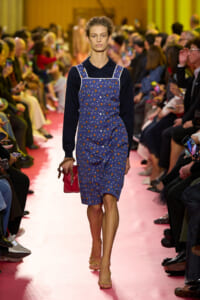 Model walking down a pink runway wearing a blue floral pinafore over a black long-sleeve top, holding a pink clutch.