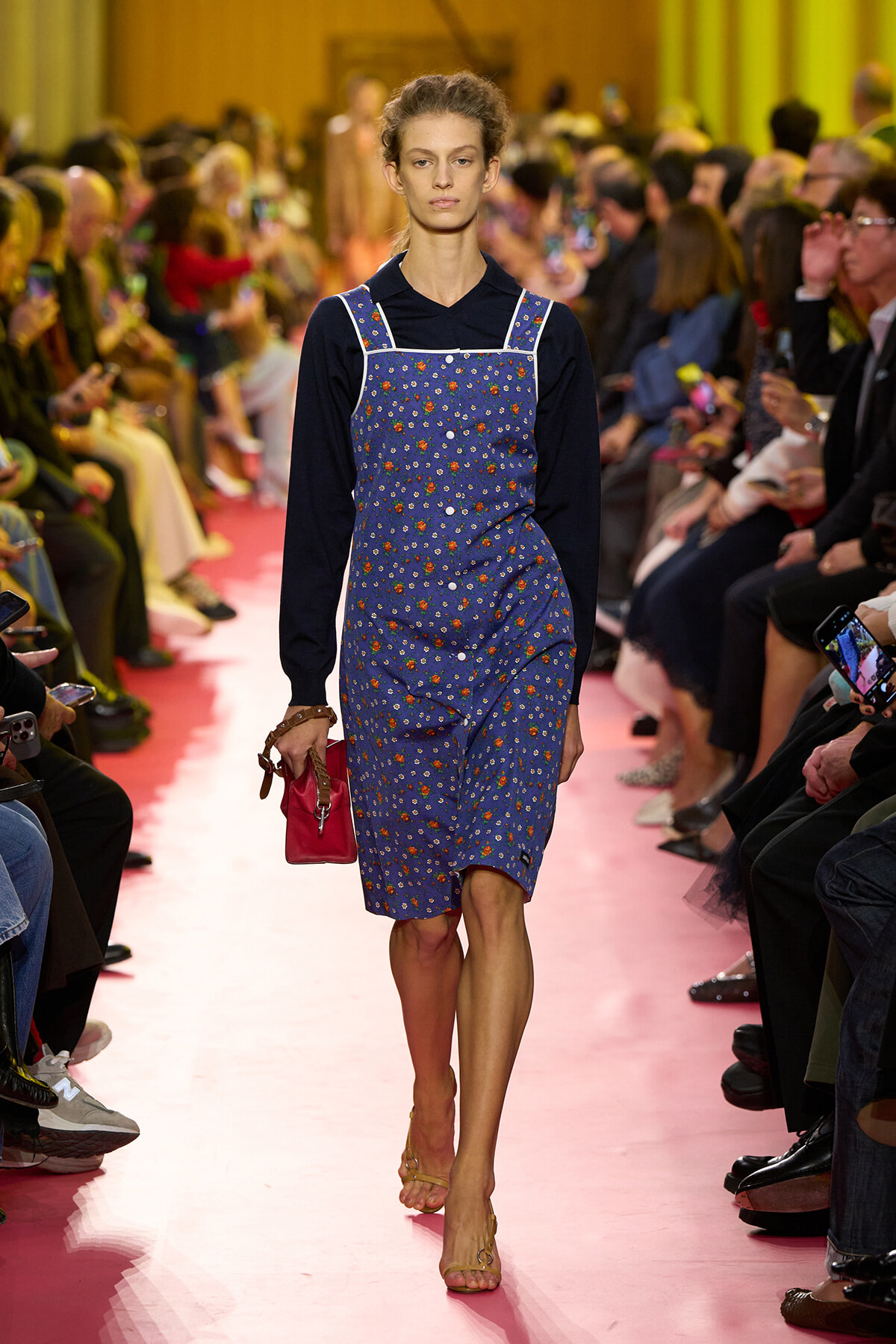 Model walking down a pink runway wearing a blue floral pinafore over a black long-sleeve top, holding a pink clutch.