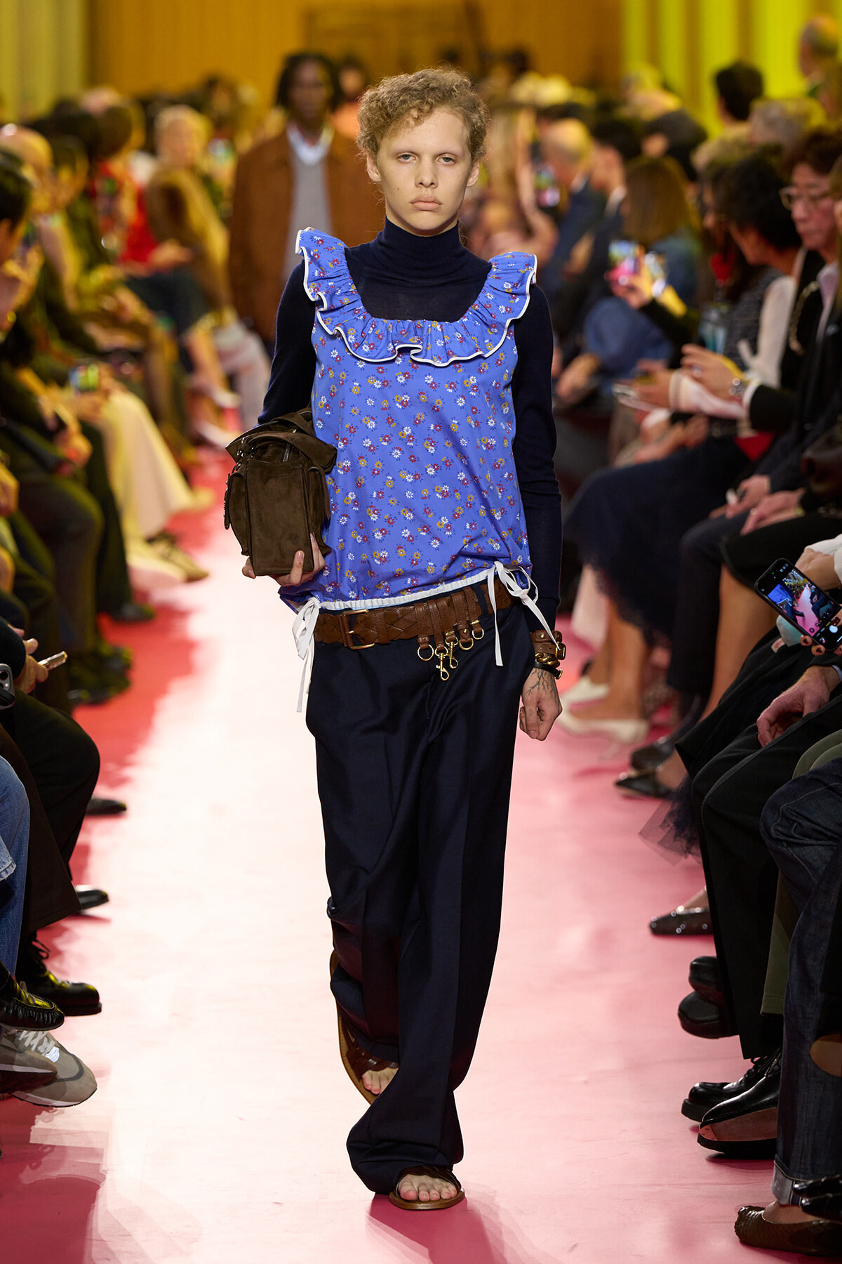 Model walks a pink runway wearing a blue floral ruffled top over a black turtleneck, dark loose pants, and sandals, with an audience in the background