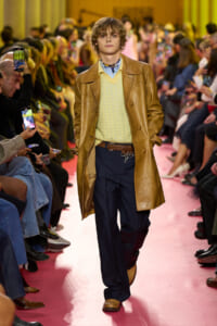 Male model walks a pink runway in a tan leather coat over a pale yellow knit sweater and navy trousers, with audience on both sides filming.