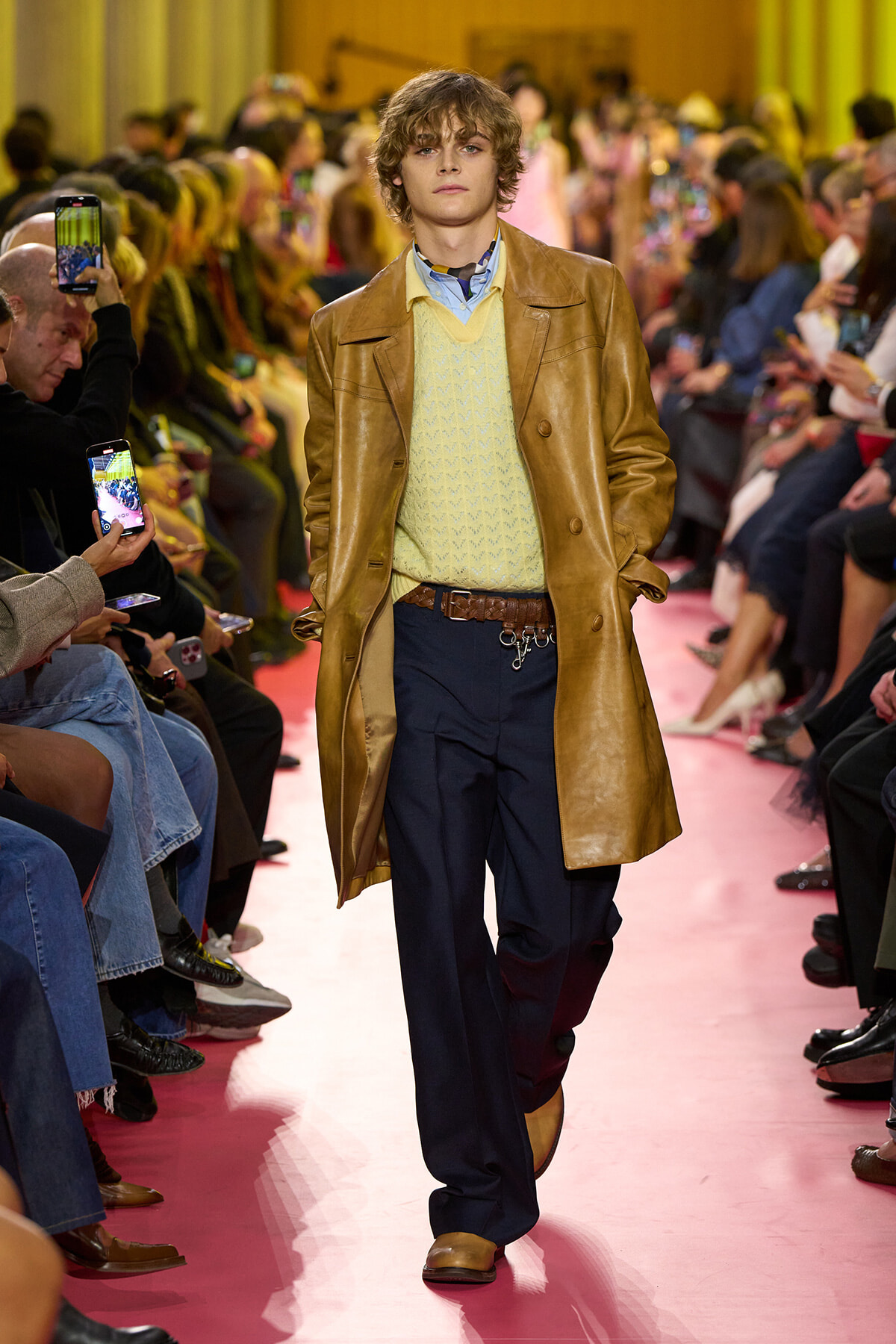 Male model walks a pink runway in a tan leather coat over a pale yellow knit sweater and navy trousers, with audience on both sides filming.