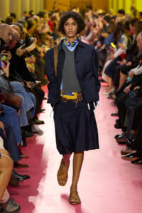 Model walking a pink runway at a fashion show, wearing a navy jacket over a gray sweater and blue shirt, with a brown belt and tan sandals while spectators watch and film.