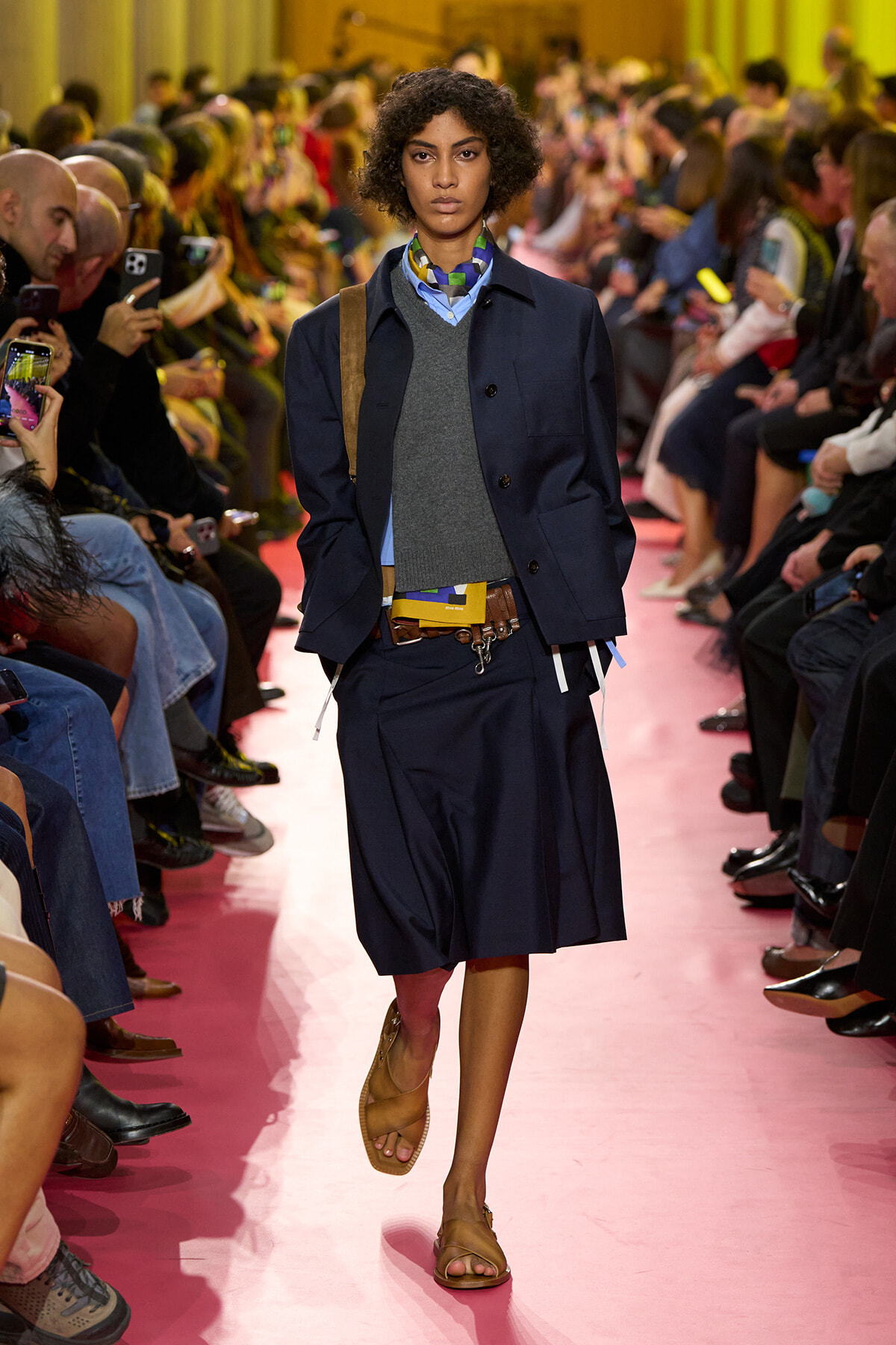 Model walking a pink runway at a fashion show, wearing a navy jacket over a gray sweater and blue shirt, with a brown belt and tan sandals while spectators watch and film.