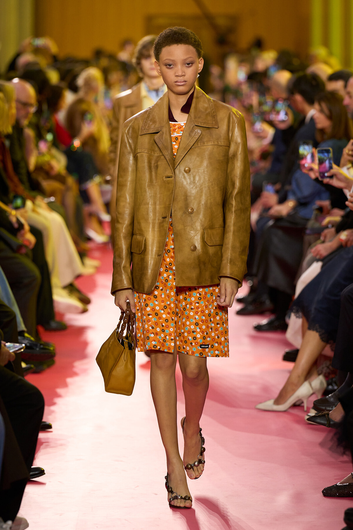 Model walking a pink runway in a brown leather blazer over an orange floral dress, carrying a tan handbag.
