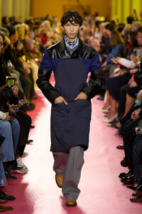 Male model struts down a pink runway wearing a navy apron over a blue jacket with black leather sleeves, gray pants, and tan shoes, crowd watching.