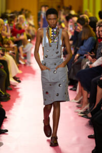 Model walking the runway in a gray sleeveless bib dress with metal studs and front pockets, pink catwalk and audience on both sides.