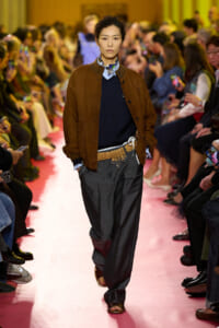 Runway model walking a pink catwalk in a brown suede jacket, navy sweater, scarf, and wide gray trousers with a braided belt and tools.