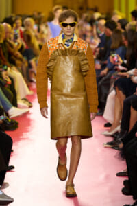 Model walks a pink runway wearing a brown leather pinafore over an orange knitted sweater and oversized yellow sunglasses, with a colorful scarf at the neck.