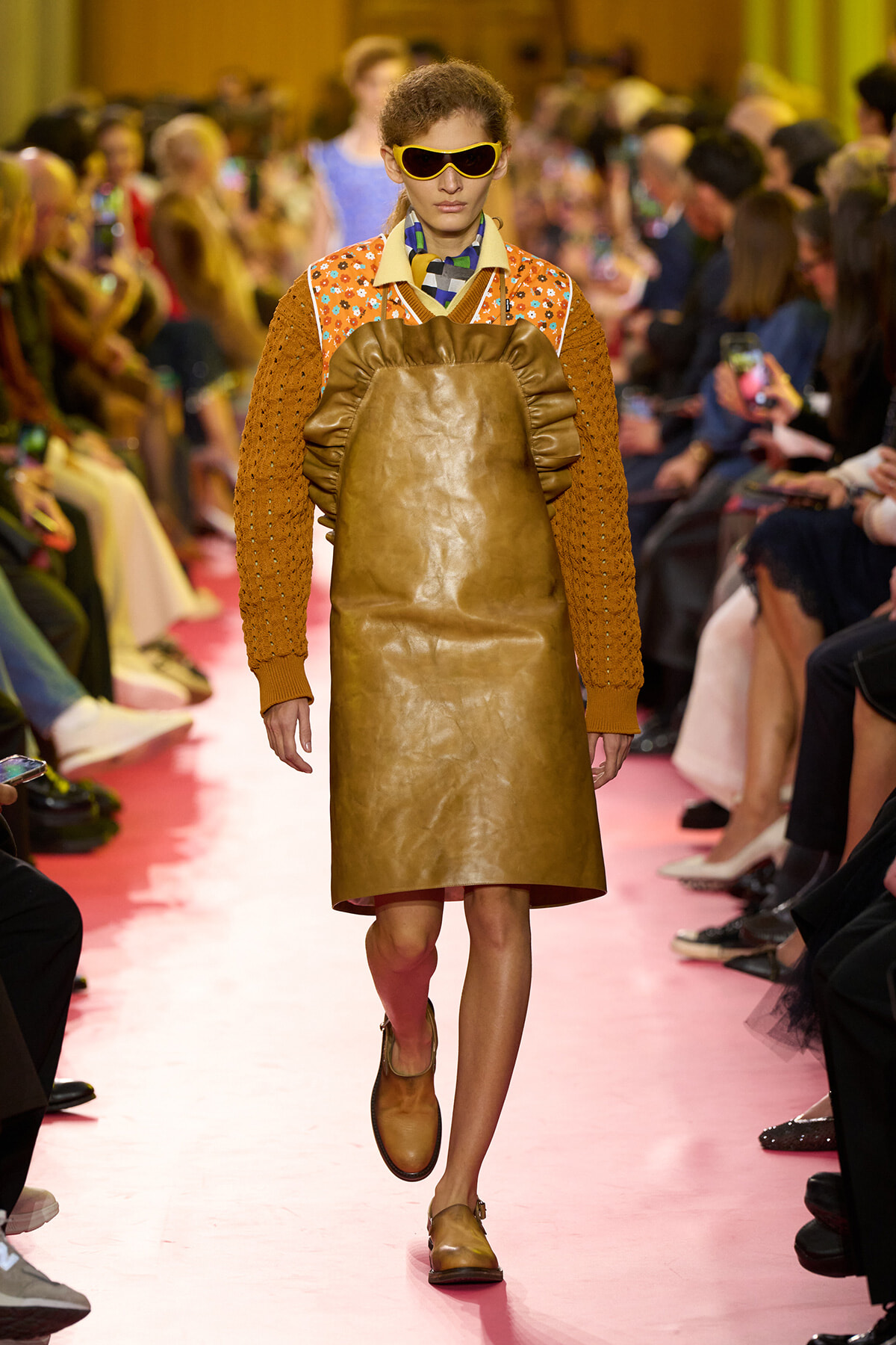 Model walks a pink runway wearing a brown leather pinafore over an orange knitted sweater and oversized yellow sunglasses, with a colorful scarf at the neck.