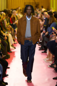 Male fashion model walks a pink runway in a brown suede jacket, grey sweater, navy pants, and a patterned scarf.