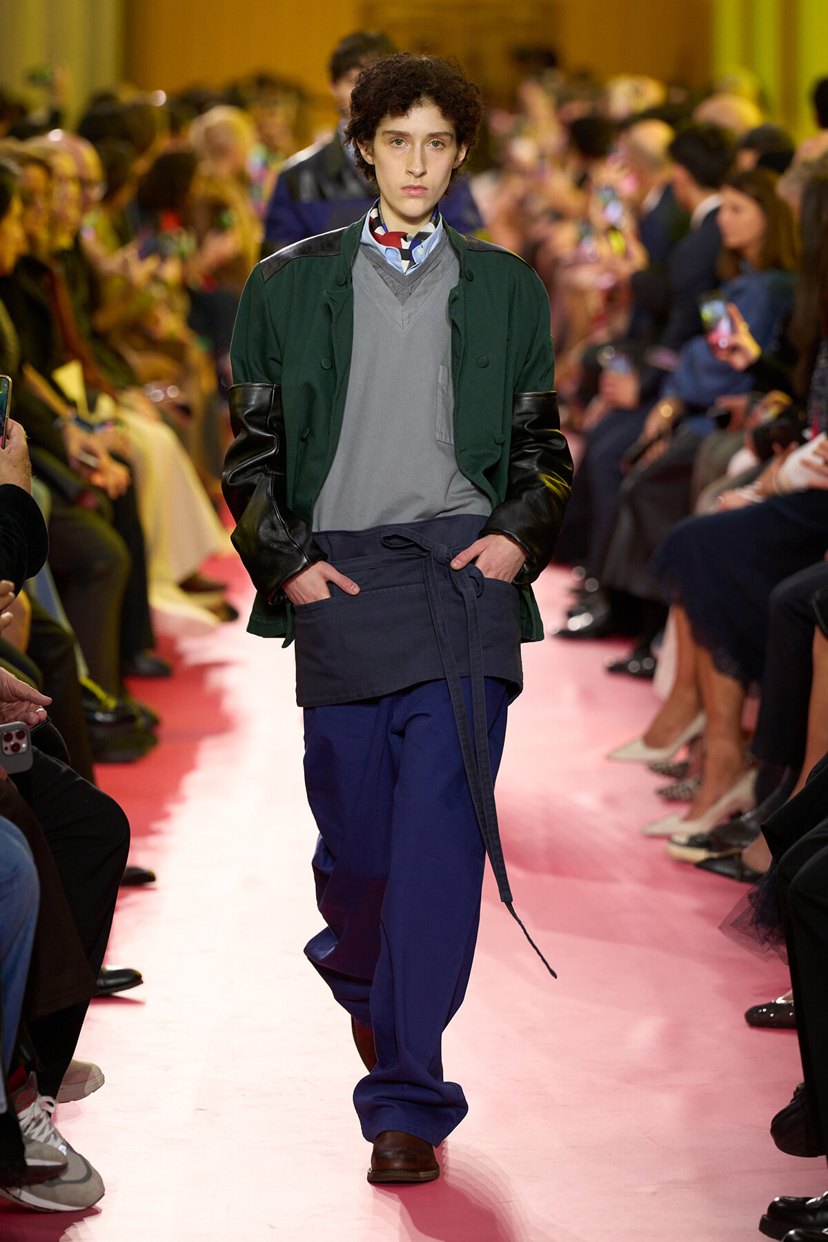 Model walks a pink runway in a dark green jacket with black leather sleeves, gray sweater, and blue pants, audience on both sides filming
