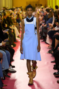 Model walks the runway in a light blue pinafore dress over a dark top, paired with tan slouch boots on a pink catwalk surrounded by spectators at a fashion show.
