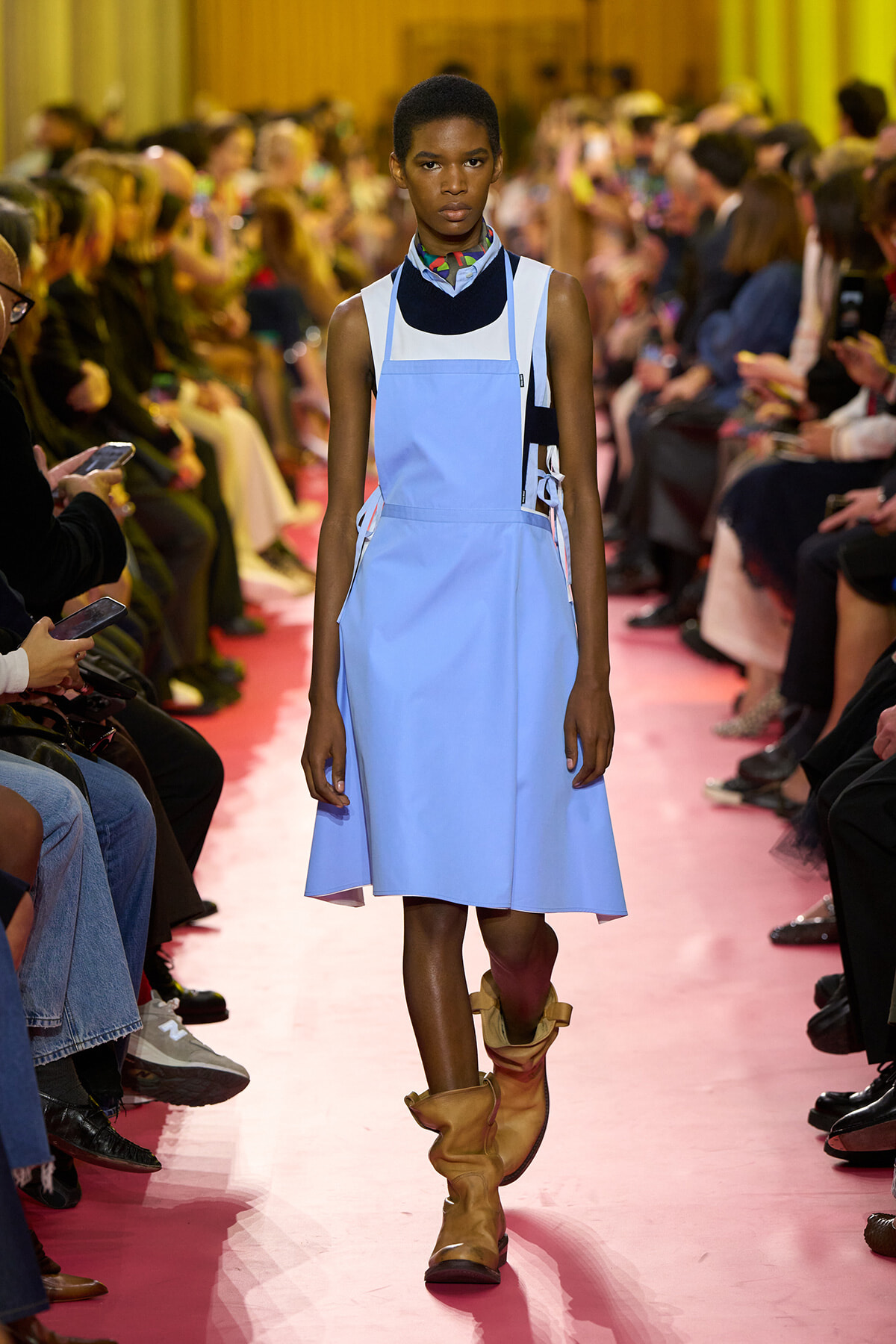 Model walks the runway in a light blue pinafore dress over a dark top, paired with tan slouch boots on a pink catwalk surrounded by spectators at a fashion show.