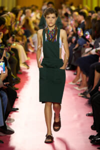 Model walking the runway in a dark green pinafore-style dress with large front pockets, layered with a light blue top and colorful scarf, as a pink catwalk and audience watch in the background.