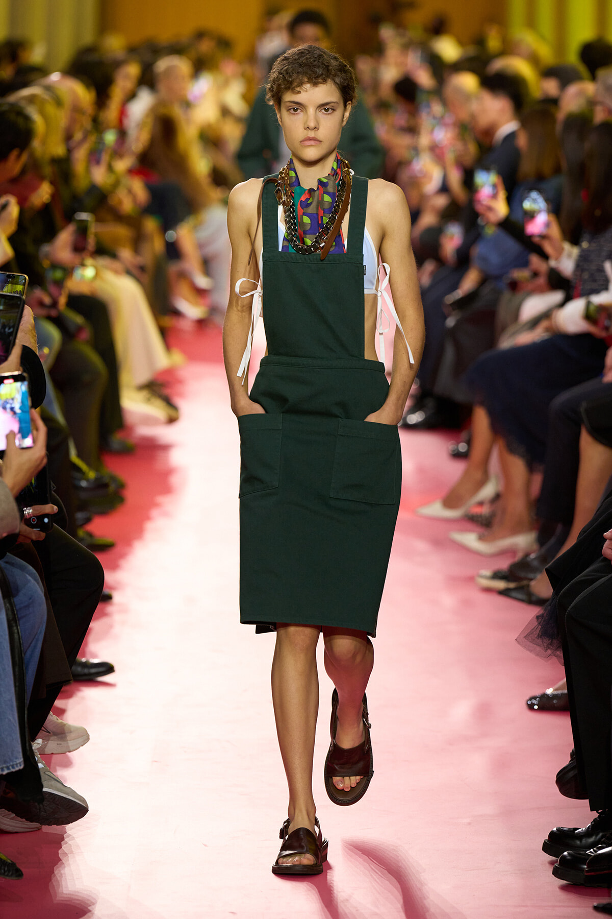 Model walking the runway in a dark green pinafore-style dress with large front pockets, layered with a light blue top and colorful scarf, as a pink catwalk and audience watch in the background.