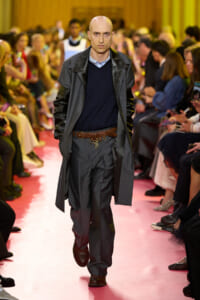 Male model walks a pink runway in a gray coat, navy sweater, and belted gray trousers at a fashion show.