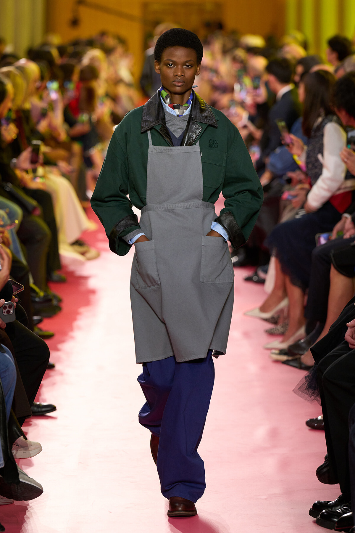 Model walks the pink runway wearing an oversized gray apron over a dark green jacket, with blue pants and hands in pockets as the audience watches.